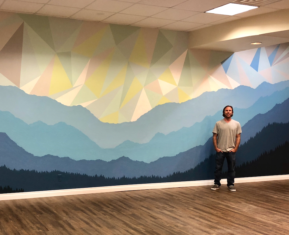 A Burnt Module mural painted in San Diego featuring an abstract triangle cloud sky design over a range of mountains. Billy Pease is standing in front of the mural that he painted.