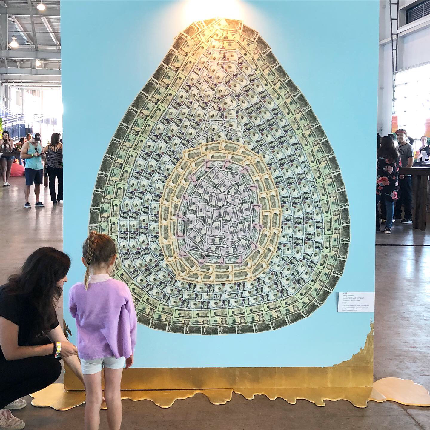 Large avocado artwork by Burnt Module  composed of paper money.  The money avocado is on a light blue background with gold leaf dripping down onto the floor.