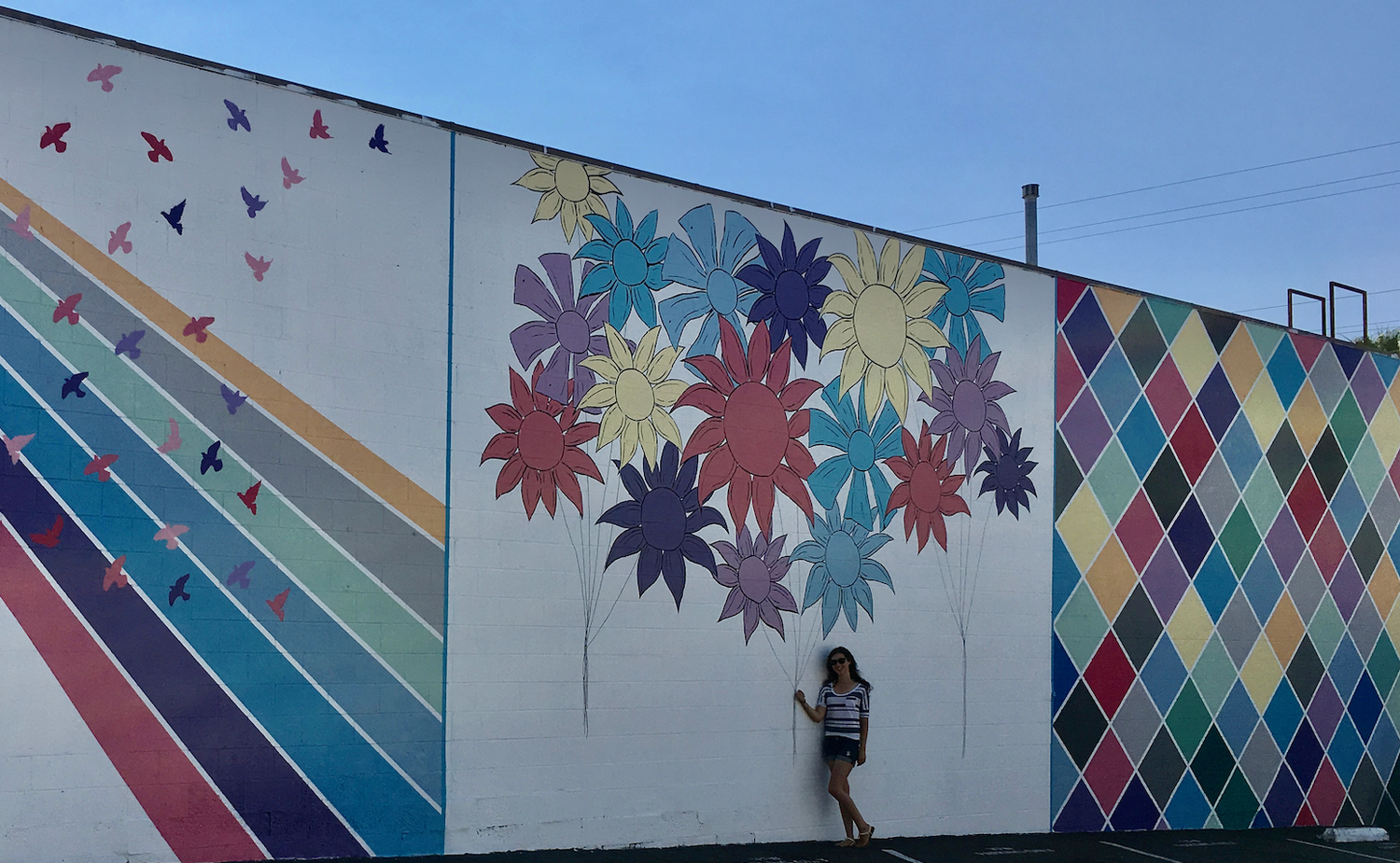 A large Burnt Module mural featuring three different sections.  The first section is rays of sun with doves flying through it.  The second section is composed of flowers attached to painted balloon strings.  The third section is a colorful square pattern.